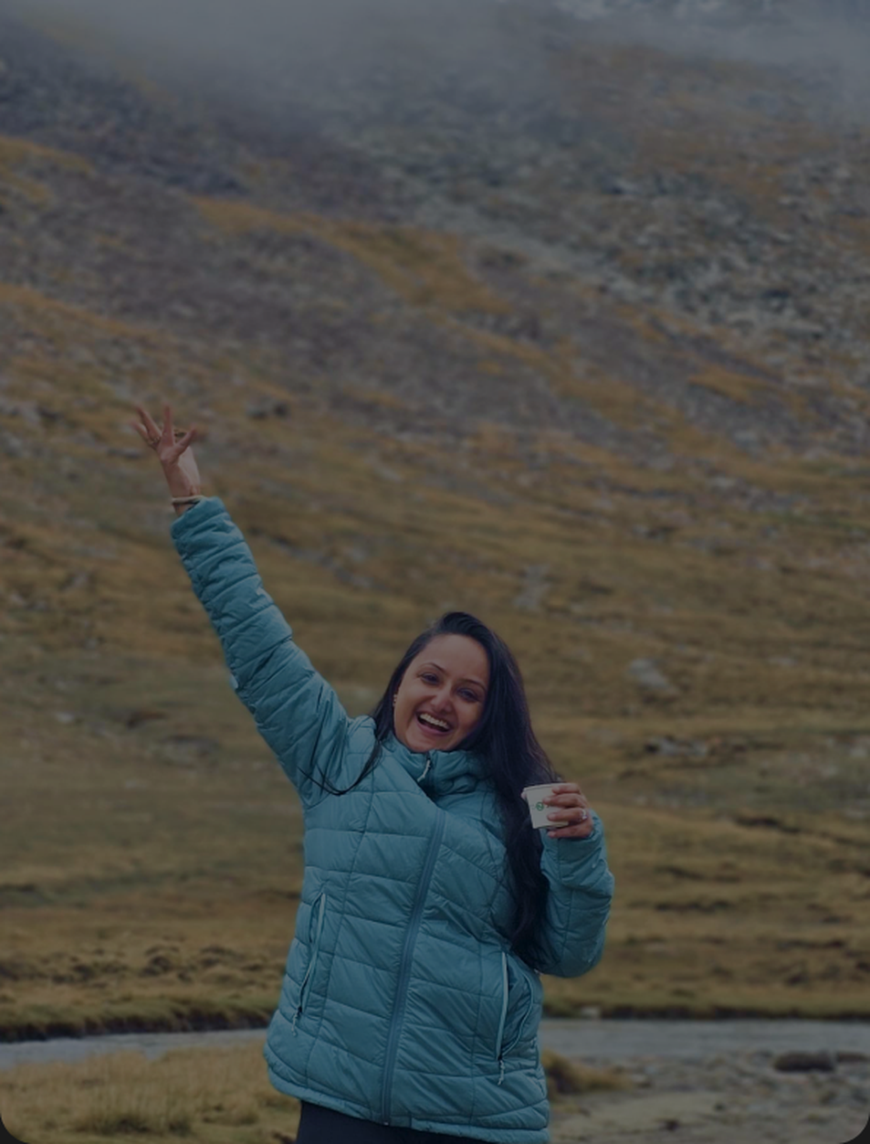 Divya on a hillside, coffee in hand, one arm up, laughing.